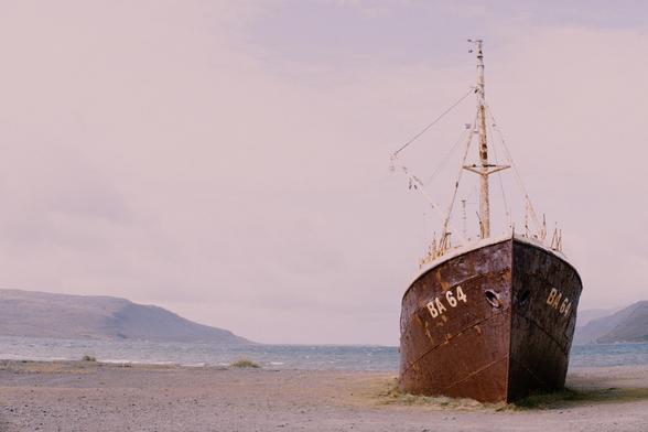 An abandoned steel ship, the Garðar BA 64, beached on a shallow sand shoreline in Iceland's Westfjords. The hull is deep red with rust, and tufts of grass have started to grow around the Norwegian built ship (1912) that was beached in 1981. Big white letters reading BA 64 can be seen near the front by the anchor holes. Some gentle slopped mountains rise up from the the horizon of the sea, against a blanket of thick, light coloured clouds.