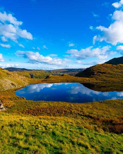 A small, still mountain lake in a valley of rolling hills covered in green, gold, and orange grasses and foliage. A bright blue sky with fluffy white clouds is reflected in the surface of the blue water.