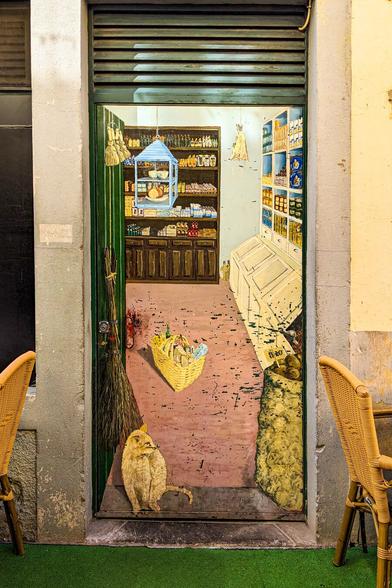A sweet street mural painted onto a door between stone walls. The artwork shows the inside of a quaint grocery store with pink floors, wooden shelves packed with goods, hanging brooms, and a wicker basket. A painted yellow cat sits on the ground looking out of the (painted) open doorway. Wicker cafe chairs sit on either side of the painted door.
