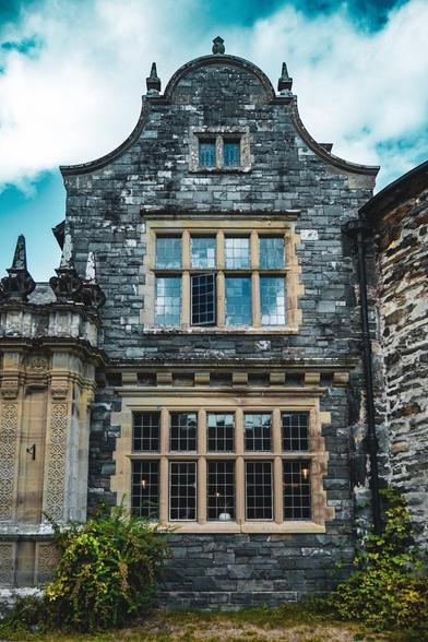 The stone exterior of a Welsh manor house featuring an open leaded glass window on the second floor, a large multi-pane window below, and lush green bushes along the base.
