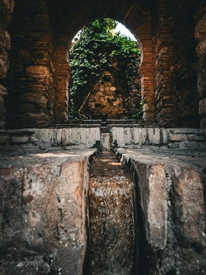 An ancient Moorish water channel with flowing water inside the Alcazaba of Málaga, a stone and brick fortress. The channel back to a semi-circular brick arch framing a stone wall partially covered with green ivy.