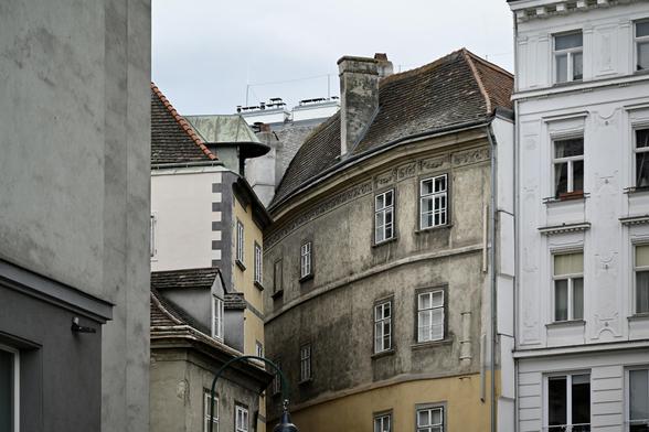 Zwischen zwei geraden Hausfassaden steht ein wenig gedrängt ein Haus mit runden Wand, die sich nach hinten wegbiegt. In der Wand weiße Sprossenfenster. Das untere der drei Stockwerke ist ockergelb geputzt, die beiden darüber sind schmutzig beige, darüber ein schwärzlich-rotes Ziegeldach.