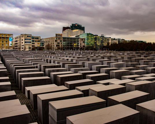 A wide-angle shot of the Memorial to the Murdered Jews of Europe in Berlin. A tight grid pattern of huge rectangular concrete blocks of differing heights spreads into the distance. In the background city buildings rise into a moody, cloudy twilight sky.