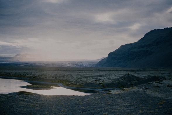 The bleak and barren landscape of northern Iceland, The sun is setting and giving the environment a eerie feeling. A dark soil/stone ground has small tufts of dark yellowish moss growing sporadically. In the distance towards the horizon, a glacier is faintly visible through an incoming haze hanging in the sky. A mountain protrudes to the right of the photo, in various shades of dark greys.