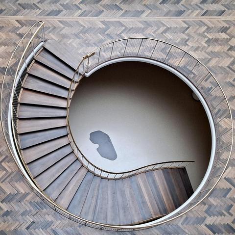 Short version: spiral staircase with foundation stone below. Fuller description: Spiral wooden staircase with brass handrail bannisters in Aarhus City Hall, Rådhuset, Denmark. Designed by Arne Jacobsen and Erik Møller. Listed building. On the floor below, the building's foundation stone. Unseen: a time capsule. When the foundation stone was laid in 1938, a time capsule was buried under it. The capsule is a lead tube containing coins from that time, newspapers of the day and a document about the start of construction. The foundation stone is black granite from the site where the city’s first town hall stood beside Domkirken (the cathedral).