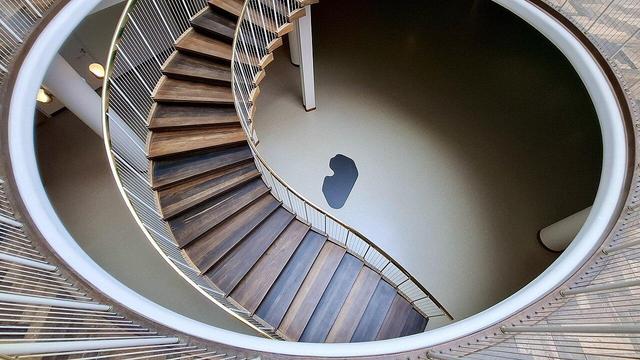 Same spiral staircase with foundation stone below, different angle. Same full Alt Text as other photos follows: Spiral wooden staircase with brass handrail bannisters in Aarhus City Hall, Rådhuset, Denmark. Designed by Arne Jacobsen and Erik Møller. Listed building. On the floor below, the building's foundation stone. Unseen: a time capsule. When the foundation stone was laid in 1938, a time capsule was buried under it. The capsule is a lead tube containing coins from that time, newspapers of the day and a document about the start of construction. The foundation stone is black granite from the site where the city’s first town hall stood beside Domkirken (the cathedral).