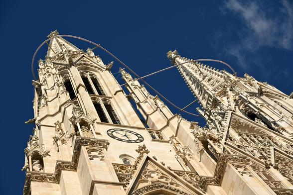 Blick von unten auf die neugotischen Türme der Votivkirche vor intensiv blauem Himmel. Um die Türme ist ein Lichtband in Form einer liegenden Acht angebracht.