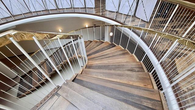 Same spiral staircase with foundation stone below, different angle. Same full Alt Text as other photos follows: Spiral wooden staircase with brass handrail bannisters in Aarhus City Hall, Rådhuset, Denmark. Designed by Arne Jacobsen and Erik Møller. Listed building. On the floor below, the building's foundation stone. Unseen: a time capsule. When the foundation stone was laid in 1938, a time capsule was buried under it. The capsule is a lead tube containing coins from that time, newspapers of the day and a document about the start of construction. The foundation stone is black granite from the site where the city’s first town hall stood beside Domkirken (the cathedral).