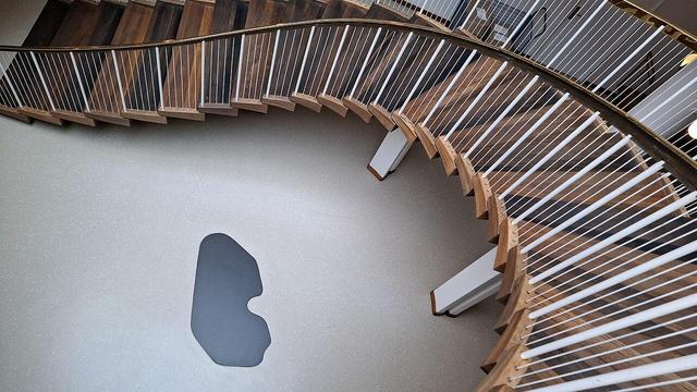 Same spiral staircase with foundation stone below, different angle. Same full Alt Text as other photos follows: Spiral wooden staircase with brass handrail bannisters in Aarhus City Hall, Rådhuset, Denmark. Designed by Arne Jacobsen and Erik Møller. Listed building. On the floor below, the building's foundation stone. Unseen: a time capsule. When the foundation stone was laid in 1938, a time capsule was buried under it. The capsule is a lead tube containing coins from that time, newspapers of the day and a document about the start of construction. The foundation stone is black granite from the site where the city’s first town hall stood beside Domkirken (the cathedral).