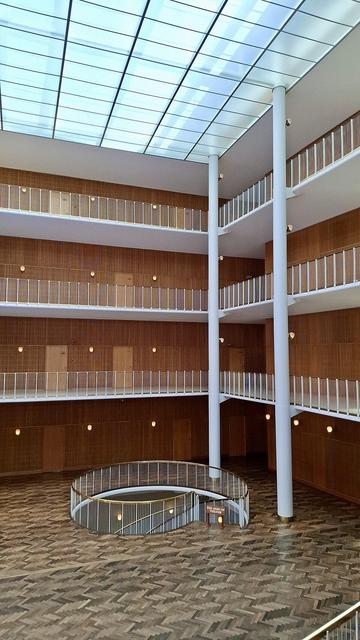 Top of spiral wooden staircase in foyer. Three upper floors above with white railings on open walkways. White columns join them. Skylights above.