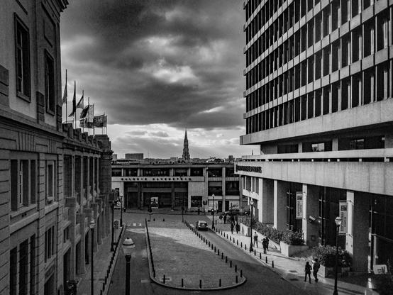 A high-angle monochrome view looking down Rue Ravenstein in Brussels. The street is framed by the historic art deco Palais des Beaux-Arts on the left and a stark modernist office building on the right. Straight ahead is the Galerie Ravenstein with the gothic spire of Brussels Town Hall visible in the far distance under a moody sky.