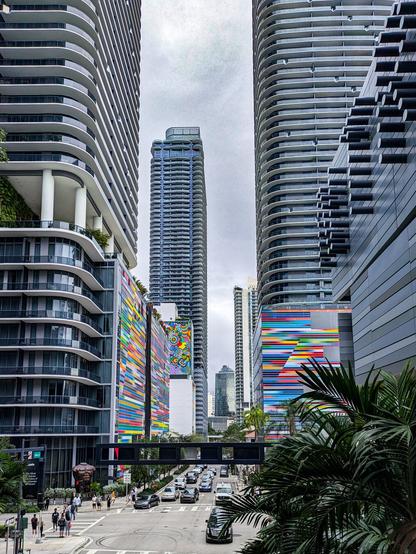 A high-angle street view of a busy intersection framed tightly on both sides by the sleek, futuristic modern high-rises with curved white balconies. The bases of the buildings feature large, vibrant public art installations in rainbow colours. Two with bold horizontal stripes and another further back with doodle art. Down the street a third similarly modern high-rise can be seen in the distance. In the foreground on the right are the dark green fronds of the top of a palm tree and in the background the sky is overcast and mostly colourless which makes the rainbow art pop in the image.