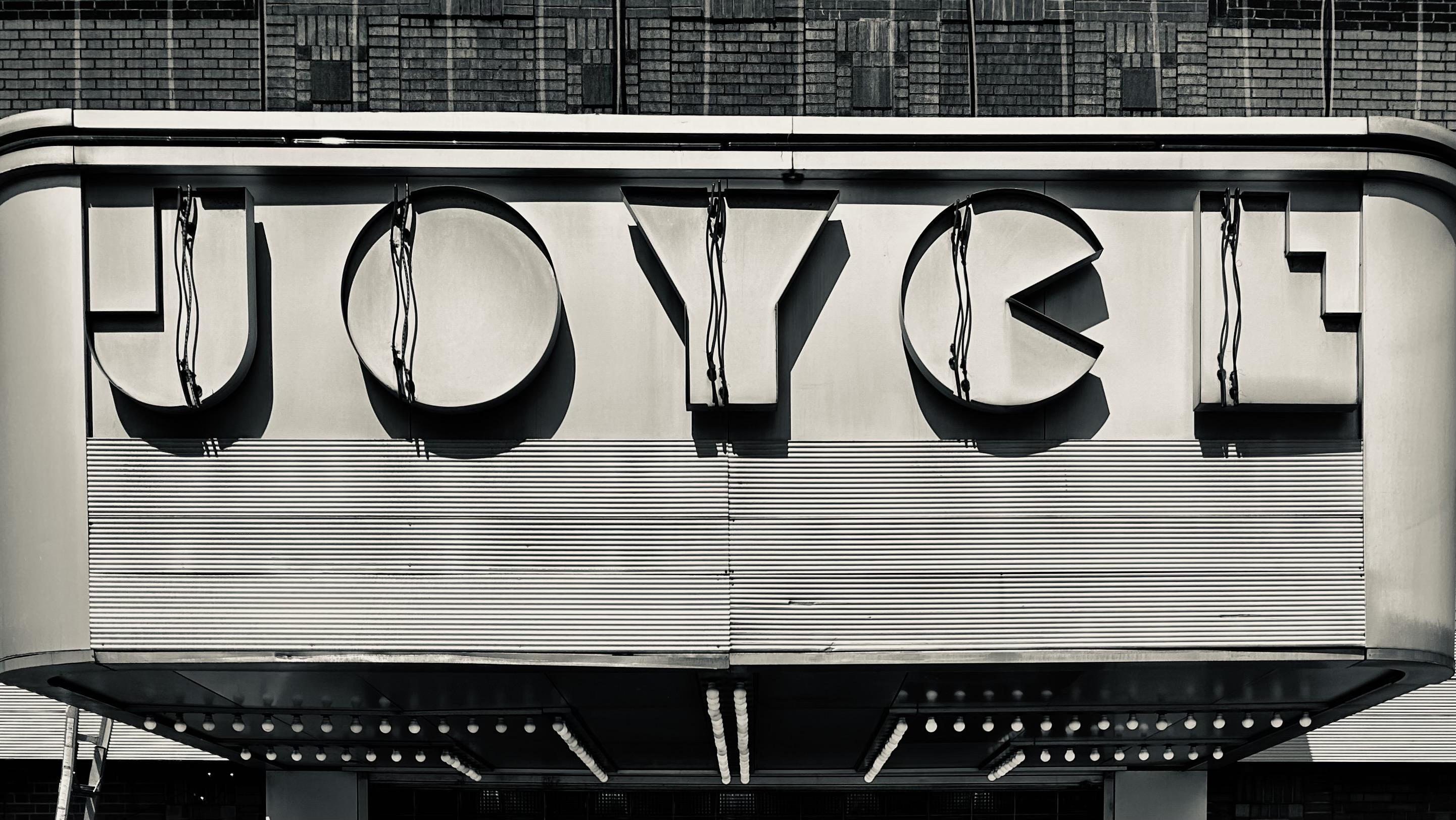 A photograph of the facade above the entrance to the Joyce Theater. It is day time, and the J O Y C E lettering is not illuminated. A ladder is leaning against the left side. Small circular lightbulbs line the underneath side leading to the entrance doors.