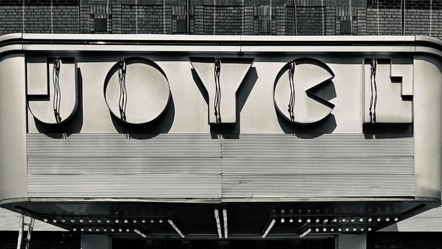 A photograph of the facade above the entrance to the Joyce Theater. It is day time, and the J O Y C E lettering is not illuminated. A ladder is leaning against the left side. Small circular lightbulbs line the underneath side leading to the entrance doors.
