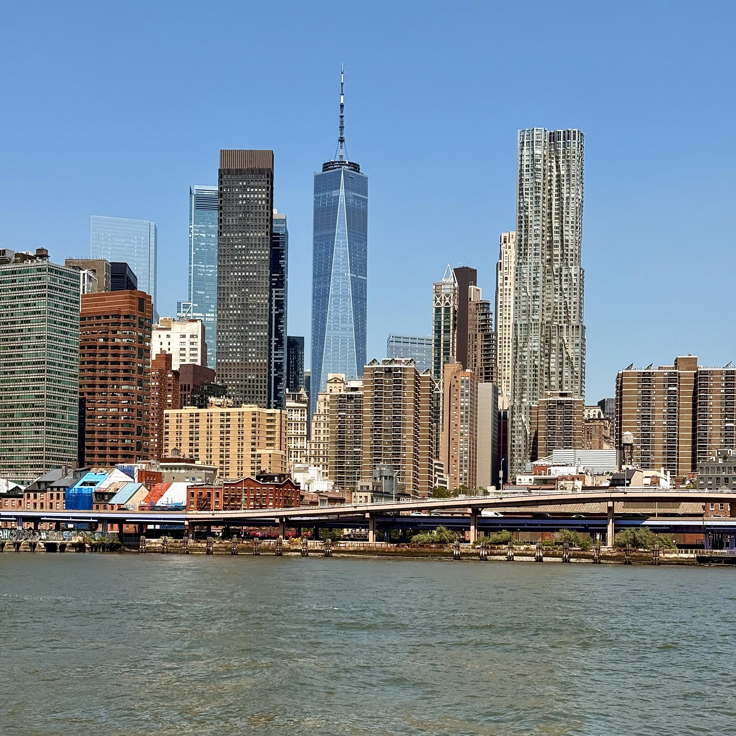 A panoramic photograph of the Manhattan skyline on a clear day, as seen from a ferry boat across the East River. The sky is a solid, cloudless cerulean blue. The foreground is dominated by the calm, greenish-brown water of the river, with some small ripples visible on its surface. An elevated, multi-lane highway, the Franklin D. Roosevelt East River Drive, runs horizontally across the mid-ground, supported by concrete pillars. Below the highway, on the riverbank, a series of low-rise buildings can be seen, including a dark brick building and some structures with colorful, graffiti-like murals on their sides. The main focus of the image is the dense collection of skyscrapers and residential buildings that make up the skyline. Dominating the center is One World Trade Center, a sleek, modern glass skyscraper tapering towards a sharp spire at its peak. To its right, the distinctive facade of 8 Spruce Street stands out, with its stainless steel panels creating a dynamic, wavy pattern. To the left of One World Trade Center, the tall, dark-colored 56 Leonard Street is visible, with its staggered, cantilevered glass blocks. Other notable buildings include a shorter, rectangular brown brick apartment complex in the foreground, and several glass and steel office towers of varying heights scattered throughout the cityscape. The buildings reflect the sunlight, and their details are sharp and clear.