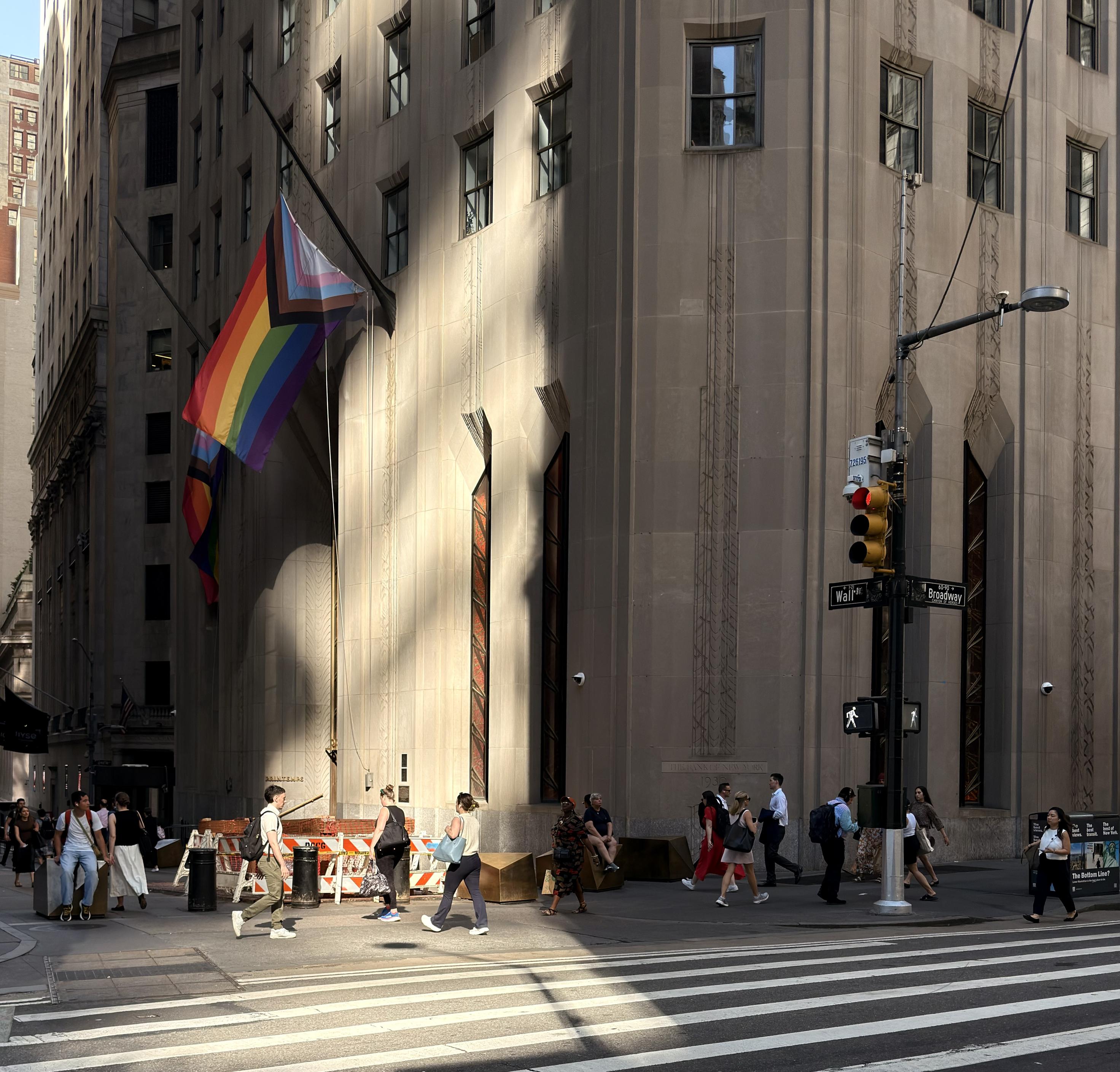 A color photograph in square aspect ratio. Taken mid morning at an intersection in the financial district of lower Manhattan. A street sign reads "Wall St" and "Broadway." The corner building on the right is a tall, stone-faced structure with two large progressive Pride flags hanging from poles. Sunlight creates a dramatic shadow on the building's facade. Pedestrians are walking in either direction on the sidewalk next to a zebra-striped crosswalk. A traffic light and street lamps are also prominent in the foreground.
