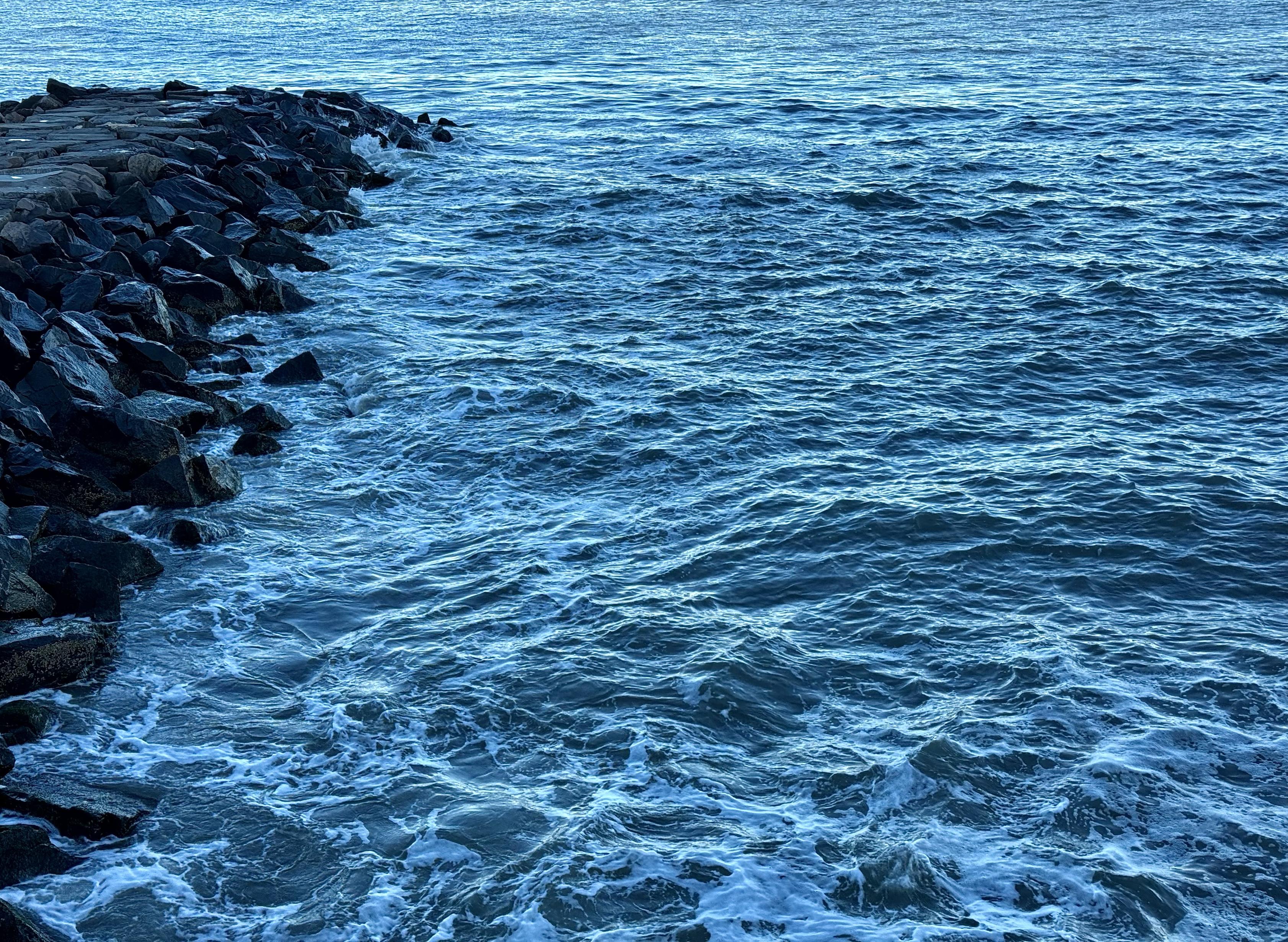 A color photograph taken mid-afternoon on a sunny day. A low-angle shot of ocean waves crashing against a rocky jetty. The water is a deep blue, with foamy white crests moving towards the shore. The rocks are dark and jagged, covered in wet moss and sea spray. The overall mood is cool and serene, with the subtle texture of the waves dominating the frame. The image is cropped so that the horizon is not visible.