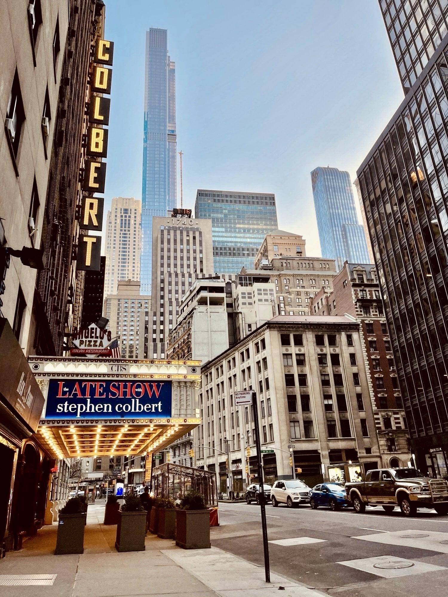 A color photograph in portrait orientation. It was taken mid morning along Broadway facing north towards West 54th street in midtown Manhattan. The focal point of the image is the Ed Sullivan Theater. Signage reads ‘The Late Show with Stephen Colbert’ in red and white lettering atop a blue background. Large, yellow illuminated letters are attached vertically to the building that read ‘Colbert’. Card are parked along the side of the street. Many tall glass skyscrapers reflecting the early morning sun are seen in the distance. A clear, light blue sky defines the horizon.