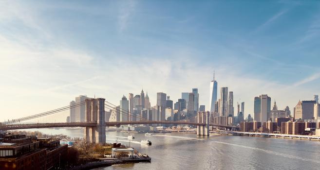 An expansive, high-angle panoramic photograph of the New York City skyline and the Brooklyn Bridge. The image is taken on a clear, sunny day, with a few wispy clouds in a bright blue sky. In the foreground, the massive stone towers and intricate steel cables of the Brooklyn Bridge dominate the left side of the frame, stretching across the East River. Below the bridge on the left bank, a small park area with bare trees and a walkway is visible, with a few small white boats in the water nearby. Brick buildings are partially visible beyond the bridge. The main focus of the image is the dense Manhattan skyline, which rises sharply from the riverbank in the midground. Iconic skyscrapers are clearly visible, including the geometric form of One World Trade Center, the slender spires of the Empire State Building and Chrysler Building, and many other modern glass and steel towers. The sunlight hits the buildings from the left, creating strong highlights and casting some shadows. The water of the East River, which separates the foreground from the midground, is calm and reflects the light, with several small boats and ferries moving across its surface.
