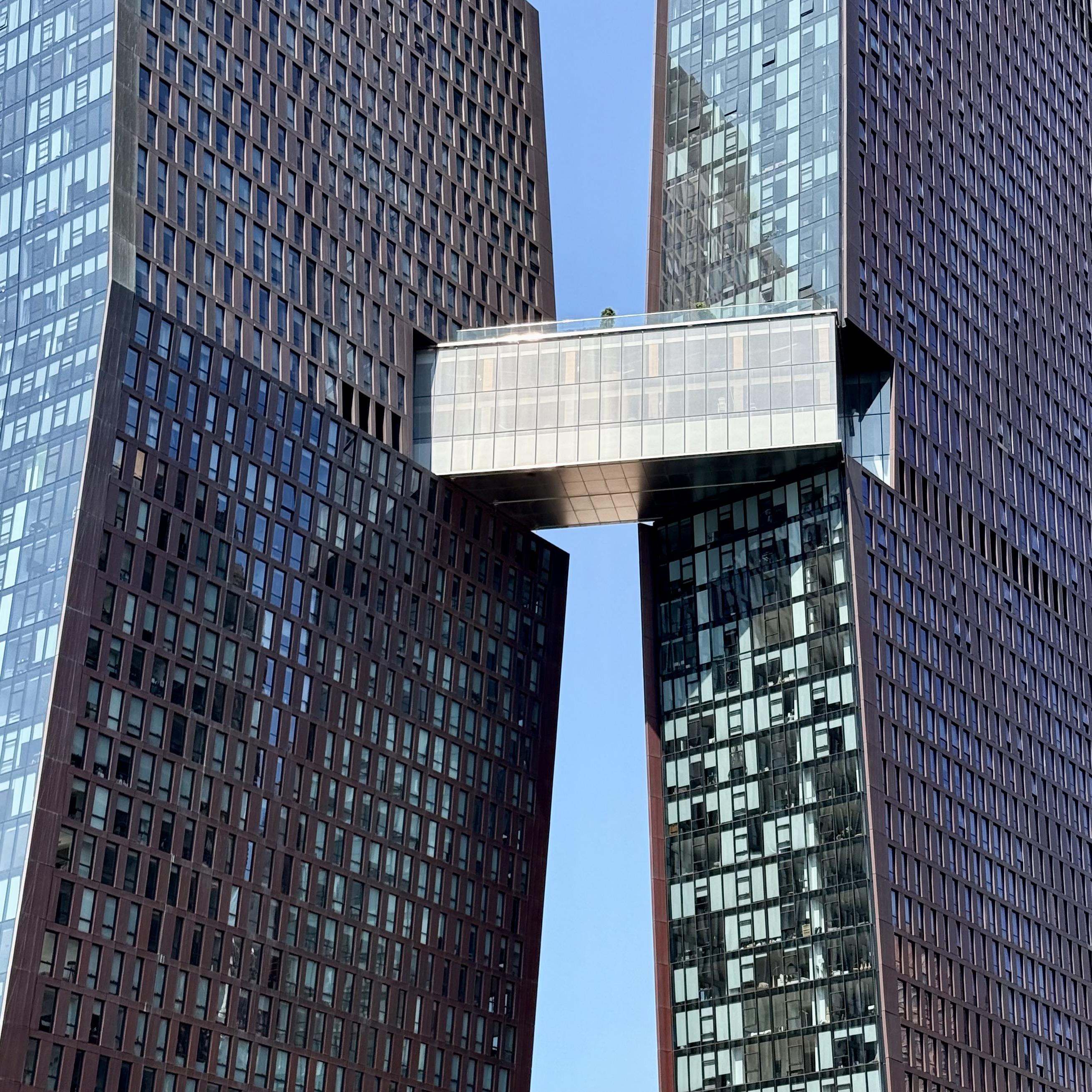 A color photograph in square aspect ratio. Taken from a low angle, showing the two tall, dark-towers of The Copper building. The towers are positioned close to each other, with a narrow gap between them that widens slightly toward the bottom. A glass-enclosed skybridge with a flat roof connects the two buildings at a high level. The windows of both towers are arranged in a regular grid pattern, reflecting the clear blue sky. The sky is bright and cloudless, and sunlight illuminates the scene, casting soft shadows. 