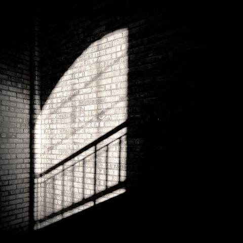 A high-contrast black and white photograph captures a bright, semi-circular spotlight of natural light from a large window outside the frame that is illuminating a corner of an old, rough brick wall. A dark, diagonal shadow cast by a handrail in the stairwell bisects the lit area. The light reveals the intricate, aged texture of the bricks, while the surrounding space is plunged into deep shadow, creating a dramatic sense of mystery and form. 