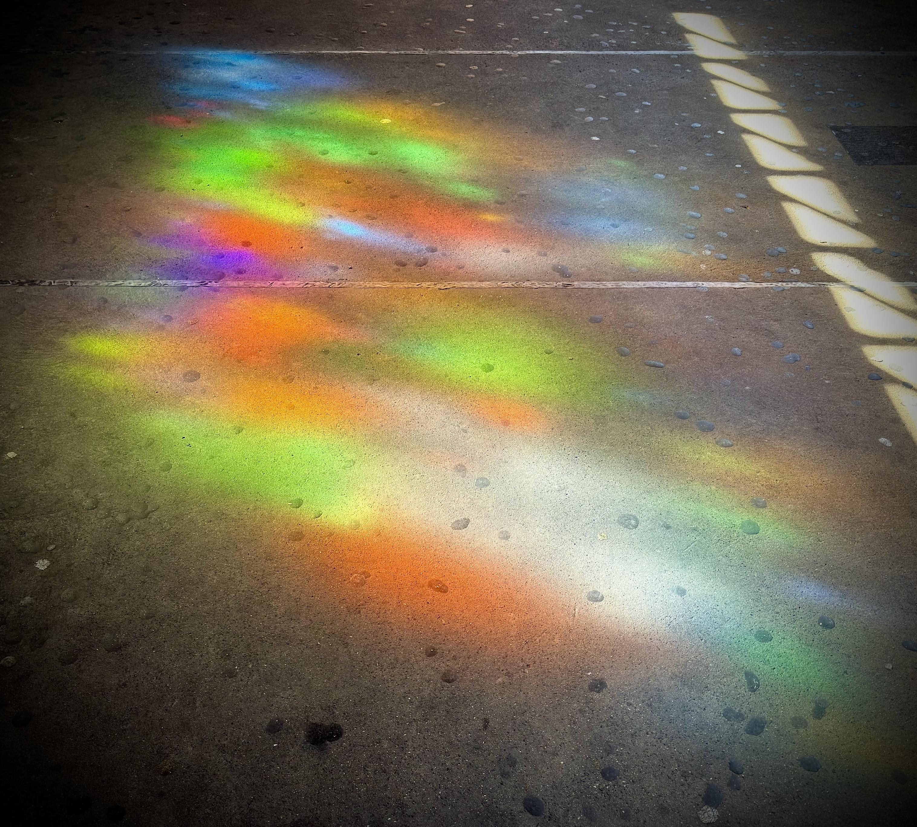 A color photograph in a square aspect ratio. The concrete platform floor at 174th street station is the focal point of the scene. Early morning sunlight is filtering through a stained glass window to the left of the camera and causing an assorted rainbow of color to cast onto the platform. A smaller set of clear windows to the left of the camera is causing a line of white light to cast onto the platform along the right edge of the image.