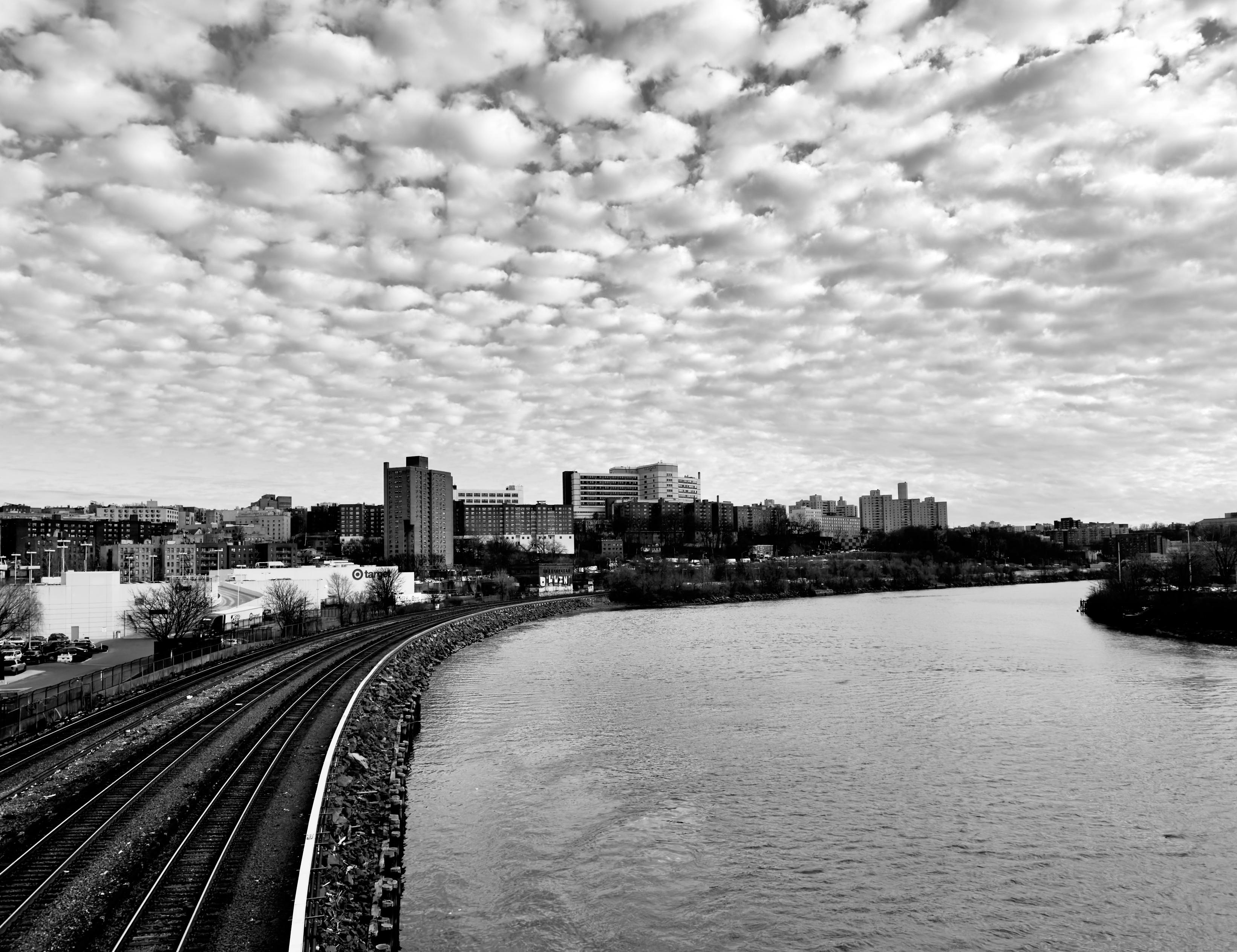 A black and white photograph taken in the Marble Hill neighborhood of Manhattan, The focal point of the image is a curved section of the Metro North train tracks running parallel to the Harlem River. The train tracks are in the foreground, bending sharply from the bottom left toward the middle of the frame before straightening out toward the right. The river occupies the majority of the lower half of the image, mirroring the curve of the tracks. Across the river and behind the tracks is the neighborhoods cityscape with various buildings, including a prominent, taller apartment building in the center, stretching across the mid-ground. The riverbank is lined with winter time trees and some industrial structures. The sky above is filled with a dense, textured layer of fluffy, altocumulus clouds filling the horizon which creates a dramatic backdrop. The overall image has a high level of contrast, emphasizing the textures of the clouds and the water.