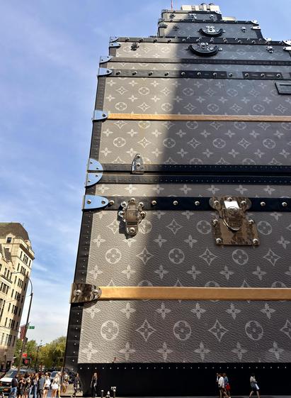 A color photograph in portrait aspect ratio. Taken along Fifth Avenue on a sunny day. The focal point of the image is the Louis Vuitton store on the corner of East 57th Street. The store has been converted to look like a gigantic stack of their luggage containers with the classic damier print, dark grey canvas with light grey insignias. Late morning sunlight is shining down onto the left side of the building, leaving the right side in shadow. On the street many people are standing and walking along the sidewalk and crossing the street. 