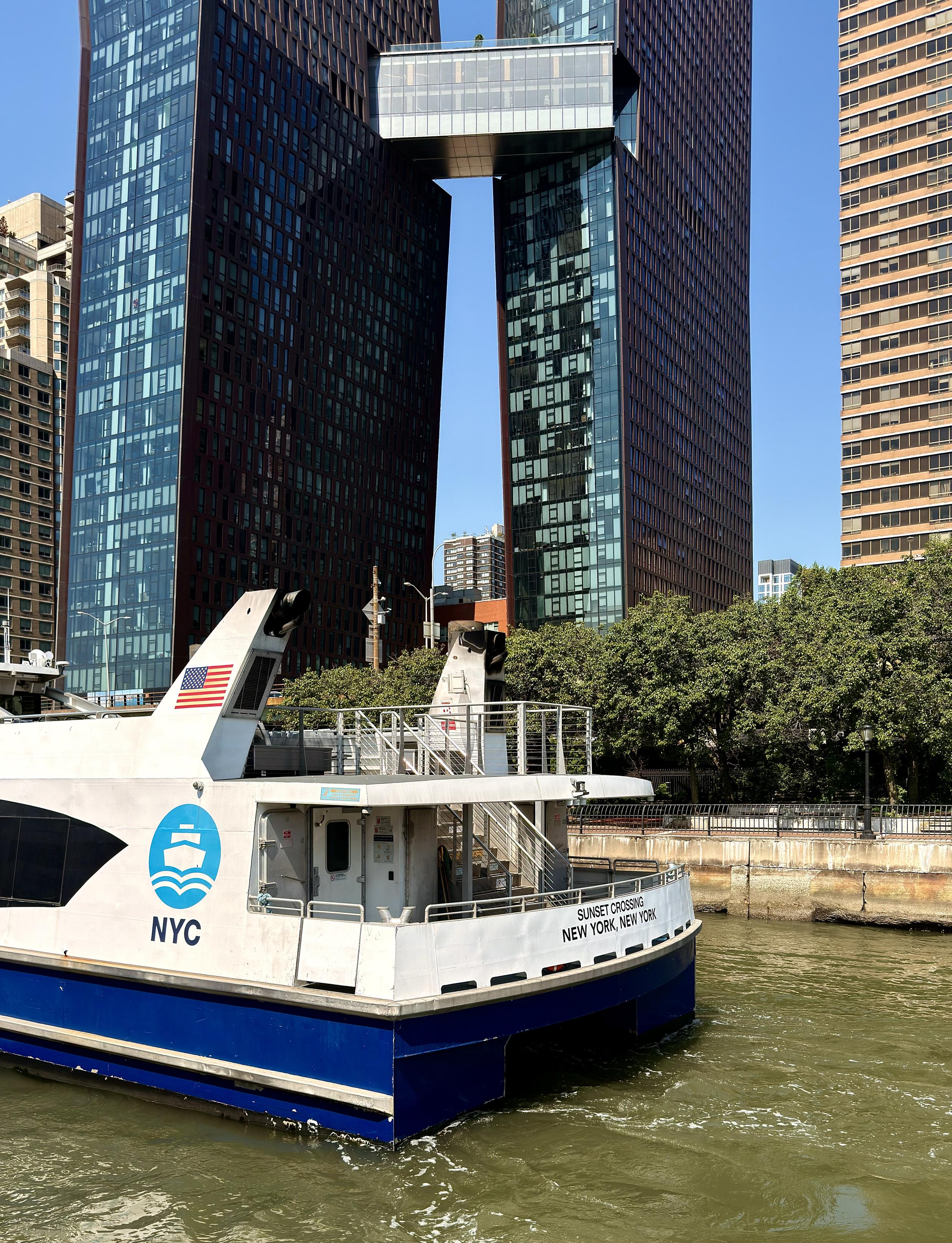 A color photograph in a portrait aspect ratio. Taken on a sunny morning from the upper deck of a ferry stopped at a dock in midtown Manhattan. The sky is bright light blue and clear. The camera is facing across the river at another ferry that is also docked in the foreground. The ferry is blue and white, with ferry signage that reads ‘NYC’ and the back of the ferry has black lettering that reads ‘Sunset Crossing New York, New York’. A large American flag adorns the top of the ferry near the stern. In the background, on the far side of a pale green colored river, is The Copper; two modern skyscrapers with dark brown, reddish-tinted glass facades. They are connected high up by a glass-enclosed pedestrian skybridge. To the right of the skyscrapers is a strip of green trees, a paved path with a black lamp post, and a taller, light-colored building in the distance.