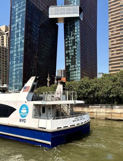 A color photograph in a portrait aspect ratio. Taken on a sunny morning from the upper deck of a ferry stopped at a dock in midtown Manhattan. The sky is bright light blue and clear. The camera is facing across the river at another ferry that is also docked in the foreground. The ferry is blue and white, with ferry signage that reads ‘NYC’ and the back of the ferry has black lettering that reads ‘Sunset Crossing New York, New York’. A large American flag adorns the top of the ferry near the stern. In the background, on the far side of a pale green colored river, is The Copper; two modern skyscrapers with dark brown, reddish-tinted glass facades. They are connected high up by a glass-enclosed pedestrian skybridge. To the right of the skyscrapers is a strip of green trees, a paved path with a black lamp post, and a taller, light-colored building in the distance.