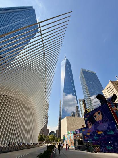A low-angle, ultra wide-shot color photograph of the buildings surrounding the transportation hub in Lower Manhattan on a clear, sunny day. The dominant feature on the left is the white, ribbed, wing-like structure of the Oculus. Its curved, skeletal form, composed of numerous parallel fins, juts out and upward, leading the eye towards the sky. To the right of the Oculus, the sleek, glass facade of One World Trade Center pierces the sky, reflecting the clouds and the surrounding buildings. In front of One World Trade Center, the low, beige-colored cube of the World Trade Center Performing Arts Center can be seen. Further to the right is 4 World Trade Center, its reflective surface mirroring the clear sky. In the foreground on the right, a vibrant, multi-colored building covered in street art, featuring a large, stylized cartoon face with wide eyes, adds a splash of color and a touch of urban grit to the otherwise clean, modern scene. The ground-level view shows a wide walkway, with a few pedestrians visible. The walkway is lined with trees and planters.