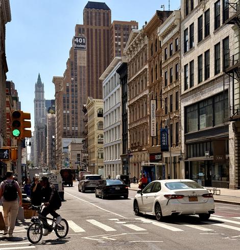 A color photograph taken on Broadway at the intersection with Grand Street in the SoHo neighborhood of Manhattan. The sky is bright and clear with no clouds. The street is lined with multi-story buildings, some with intricate architecture and fire escapes. The main focus is the view down the bustling street toward a tall, distinct green-roofed skyscraper in the distance. The street is busy with people, a bicyclist, and cars. In the foreground, a white sedan is crossing a crosswalk from right to left, and a man on a bicycle is riding toward the viewer. The green light is visible on the traffic signal. The 401 Broadway building sign is prominently displayed on one of the tall buildings as white text on a black background.