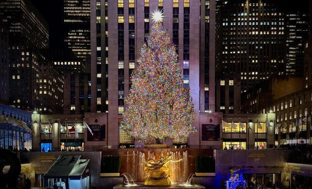 A color photograph that was shot at a wide angle, taken at nighttime of the 2024 Rockefeller Center Christmas Tree in New York City. The massive tree is brightly lit with a dense array of multicolored lights and topped with a large, glowing star. It is positioned in the center of the frame, towering over a bronze statue of Prometheus in the foreground, which is surrounded by a small fountain and illuminated from below. The surrounding buildings are visible in the background, with countless windows aglow, creating a cityscape backdrop for the main attraction. There are a few people visible at the base of the tree and along the sides of the plaza. 