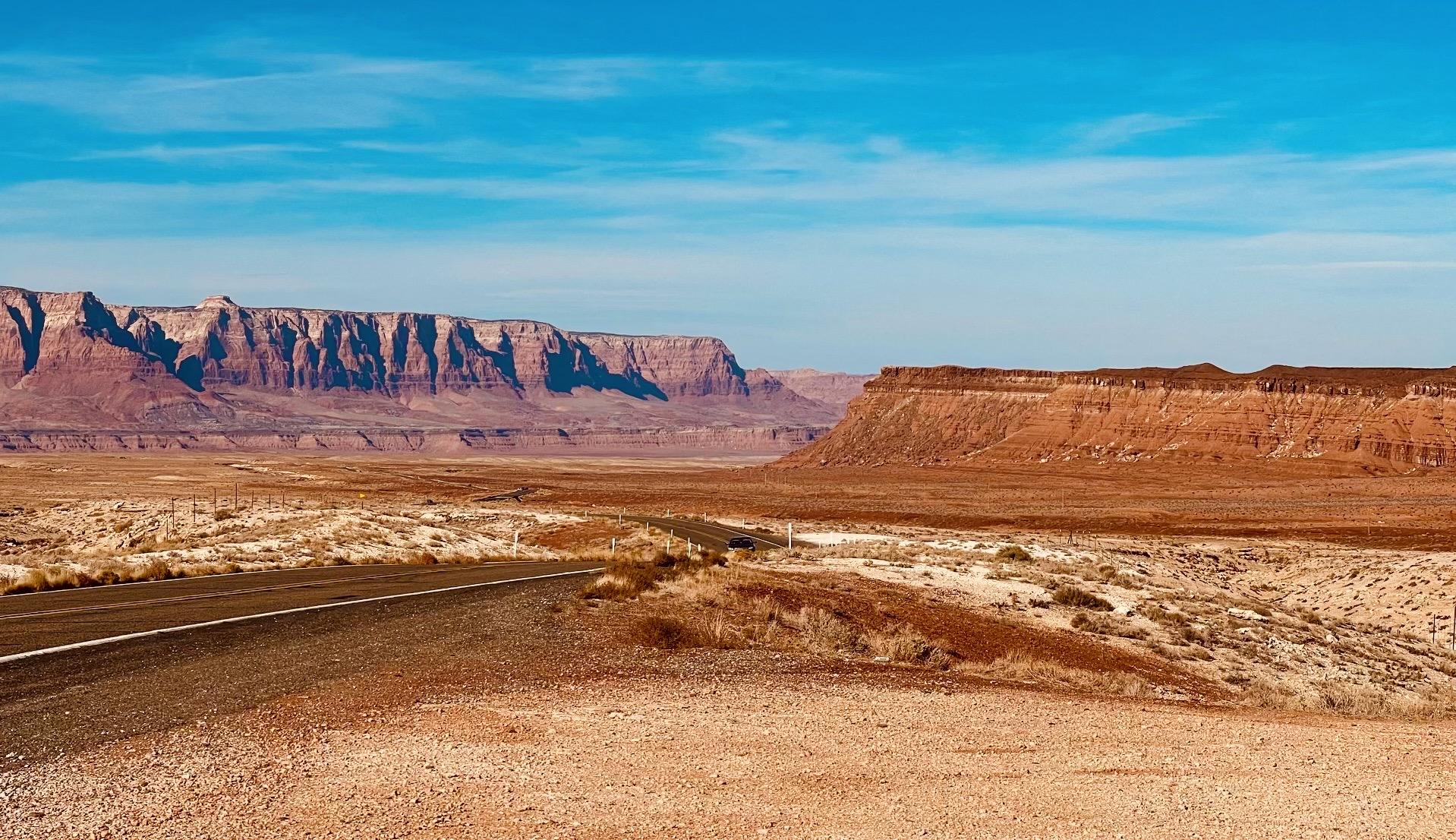 A color photograph in a landscape orientation. Two-lane highway curves through a vast, arid desert landscape. The road is dark asphalt with a white stripe down the middle, and it disappears into the distance between two massive buttes. The butte on the left is a long, layered cliff face with gray and red rock formations. On the right, the butte is a deep reddish-brown with a flatter top. The desert floor is covered in light brown and tan sand with sparse, dry brush. The sky is a vibrant, clear blue with a few wispy clouds.