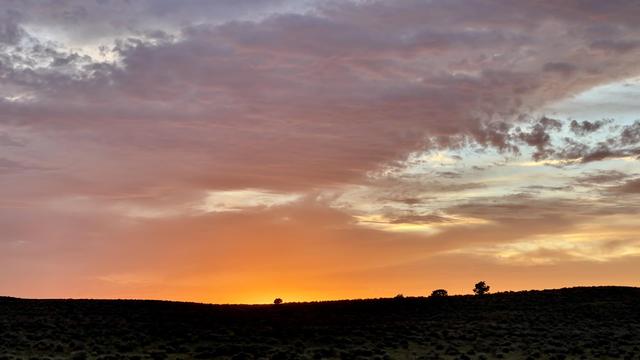 A color photograph in landscape orientation of a sunset over a low, rolling hill. The sky is a dramatic gradient of colors, from a warm, intense orange at the horizon to soft shades of purple and gray in the clouds above. The clouds themselves are textured and wispy. The hill in the foreground is a mostly dark silhouette, with only a few small trees and desert shrub bushes visible against the bright sky. 