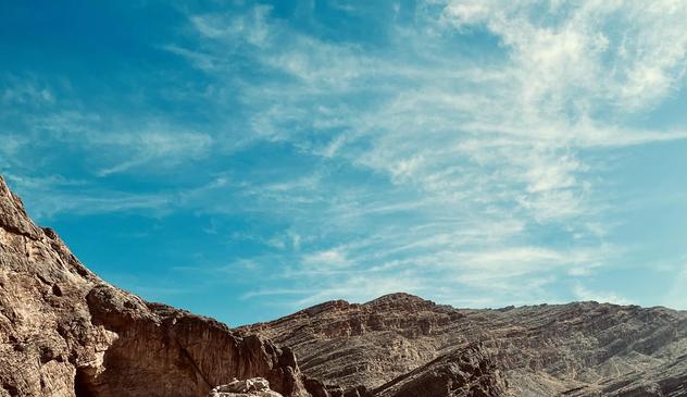 A color photograph shot at a low-angle of a desert landscape featuring two large, rocky mountain formations. On the left, a jagged, dark brown rock face is prominently featured. In the center and to the right, a wider, multi-layered mountain ridge stretches across the horizon. Above, the deep blue sky is filled with delicate, white clouds streaking across the frame.