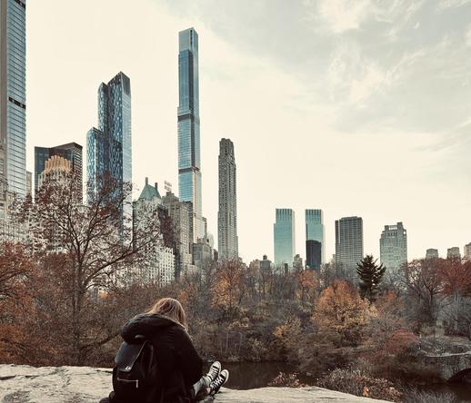 A color photograph taken near The Pond in Central Park. The focal point of the image is a person with long red hair seen from behind while sitting on a large rock overlooking the water. They are wearing a black jacket, a black backpack, and black Converse sneakers. They’re facing down reading a book that isn’t visible in the photo while their legs are crossed in front of them. Bare branches of trees in the foreground frame a view of the park's autumn foliage, with a mix of green, orange, and yellow leaves. In the distance, the midtown Manhattan skyline rises above the trees, with prominent skyscrapers like the Central Park Tower and 111 West 57th Street visible under a light cloudy sky.