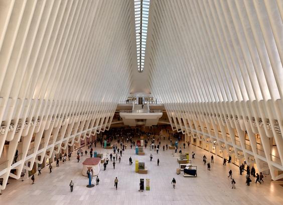 A color photograph taken from the observation deck on the upper floor of The Oculus. A symmetrical, wide-angle interior shot of the main concourse. The building's white, wing-like architecture dominates the upper half of the frame, with massive, curved steel ribs soaring upward to a central skylight, which is barely visible. The light-colored marble floor is scattered with many people, but is not busy compared to typical rush hour foot traffic. Various people are walking, standing, or sitting. Some small kiosks are present on the concourse floor. The upper levels of the structure are visible on the left and right, lined with people looking down onto the main floor. On the lower right side of the frame many bright lights from a retail outlet is illuminating the floor. Early morning light is bright and airy, reflecting off the white surfaces. 