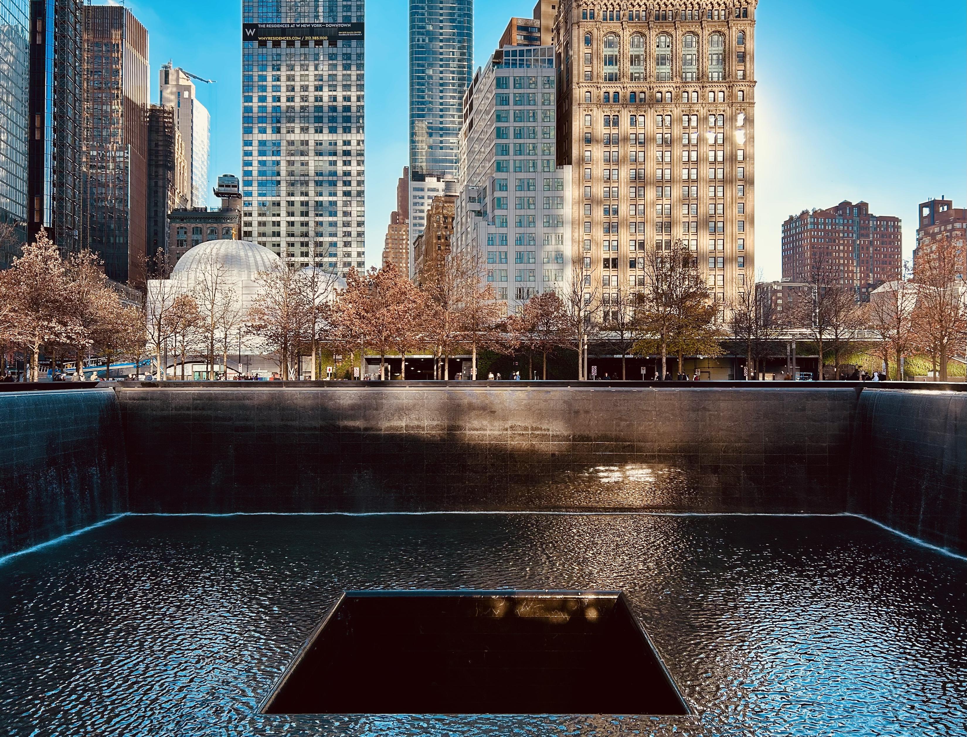 An eye-level landscape photograph of the 9/11 memorial pool in New York City on a bright, sunny day in November. The large, dark reflecting pool, framed by a cascading waterfall, takes up the foreground and a good portion of the middle ground. A smaller, square-shaped void sits within the center of the pool. The surface of the water reflects the bright blue sky and the surrounding buildings. Beyond the pool, a line of deciduous trees with bare branches and a few remaining brown leaves extends across the middle ground. Pedestrians can be seen walking along the plaza behind the trees. The background is filled with a dense collection of towering skyscrapers under a clear blue sky, including the residential skyscraper at 101 Warren Street and the Woolworth Building's recognizable architectural facade. The sunlight creates a strong contrast between the dark surface of the memorial pool and the brightly lit buildings.