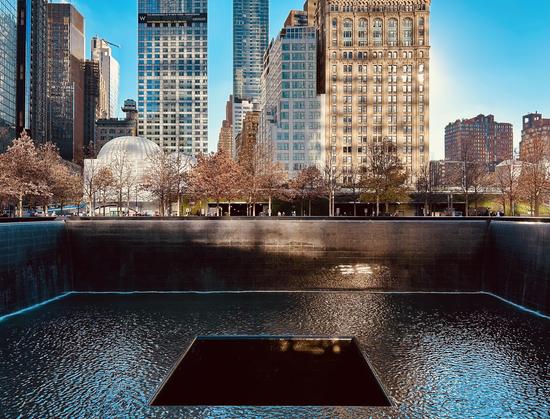 An eye-level landscape photograph of the 9/11 memorial pool in New York City on a bright, sunny day in November. The large, dark reflecting pool, framed by a cascading waterfall, takes up the foreground and a good portion of the middle ground. A smaller, square-shaped void sits within the center of the pool. The surface of the water reflects the bright blue sky and the surrounding buildings. Beyond the pool, a line of deciduous trees with bare branches and a few remaining brown leaves extends across the middle ground. Pedestrians can be seen walking along the plaza behind the trees. The background is filled with a dense collection of towering skyscrapers under a clear blue sky, including the residential skyscraper at 101 Warren Street and the Woolworth Building's recognizable architectural facade. The sunlight creates a strong contrast between the dark surface of the memorial pool and the brightly lit buildings.