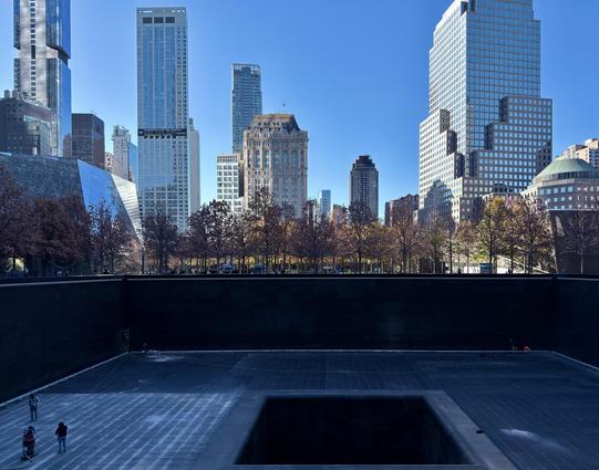 A color photograph taken as a wide-angle shot of the 9/11 Memorial & Museum in New York City on a crisp, bright late autumn day. The sky is a deep, cloudless blue. In the foreground is the memorial pool, a large, dark-colored square with a central, cascading waterfall section. The pool has been drained of water so that it can be cleaned. Several maintenance workers can be seen standing and walking in the vast cavity of the pool. In the background, beyond the memorial's plaza, is a row of trees with sparse, reddish-brown and orange foliage, typical of late autumn. Dominating the mid- and background are several prominent skyscrapers under a clear blue sky. From left to right: the reflective, angular glass facade of the One World Trade Center museum entrance is partially visible; a very tall, slender skyscraper with a geometric pattern of windows; the distinctive, ornate brown building with a pointed top, which is 22 Cortlandt Street; and the broad, terraced facade of Brookfield Place on the right, with its dome visible behind the terrace. Sunlight is reflecting off of the buildings and and the pool is in shadow.