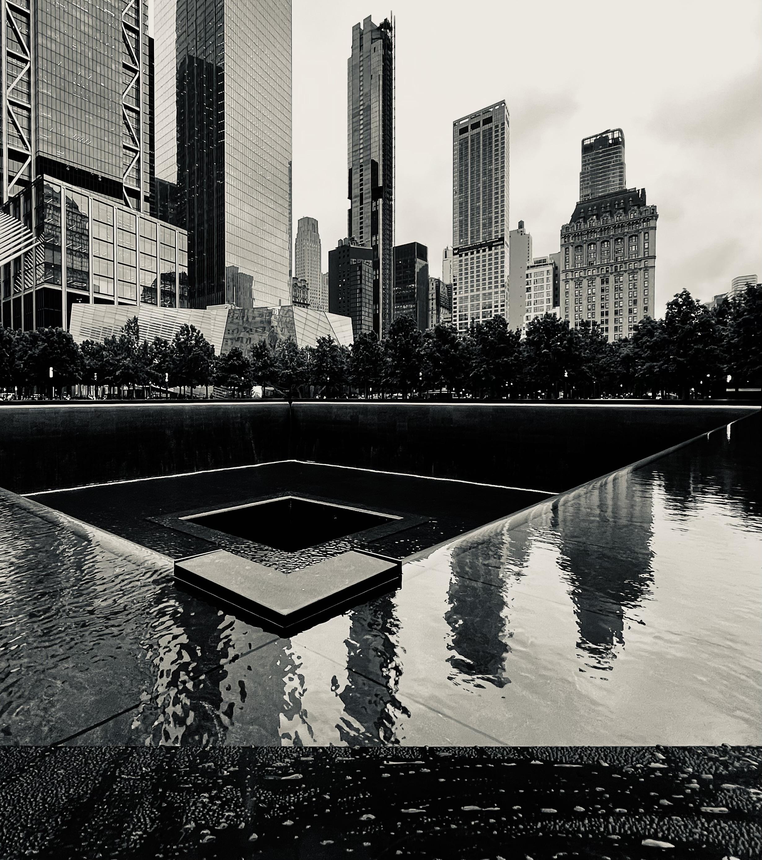 A black and white, wide-angle photograph of the 9/11 Memorial's reflection pool in New York City. The image is taken from a low angle, looking towards the northern edge of the pool. The massive, dark bronze parapet, which features the names of victims, forms a dark, sharp line across the middle ground. Water cascades down the sides of the parapet into a lower basin, which is not visible. The surface of the pool, which is flat and reflective, fills the bottom half of the frame. The water is textured with small ripples, reflecting the surrounding skyscrapers and the cloudy sky. In the background, the modern skyline of Lower Manhattan is visible. The buildings are a mix of glass and steel. The sky above is overcast, creating a muted light that enhances the somber mood of the scene. A line of trees stands between the pool and the buildings, adding a natural element to the otherwise urban landscape. The overall tone is one of quiet contemplation and solemnity.
