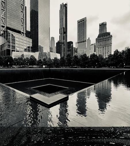A black and white, wide-angle photograph of the 9/11 Memorial's reflection pool in New York City. The image is taken from a low angle, looking towards the northern edge of the pool. The massive, dark bronze parapet, which features the names of victims, forms a dark, sharp line across the middle ground. Water cascades down the sides of the parapet into a lower basin, which is not visible. The surface of the pool, which is flat and reflective, fills the bottom half of the frame. The water is textured with small ripples, reflecting the surrounding skyscrapers and the cloudy sky. In the background, the modern skyline of Lower Manhattan is visible. The buildings are a mix of glass and steel. The sky above is overcast, creating a muted light that enhances the somber mood of the scene. A line of trees stands between the pool and the buildings, adding a natural element to the otherwise urban landscape. The overall tone is one of quiet contemplation and solemnity.