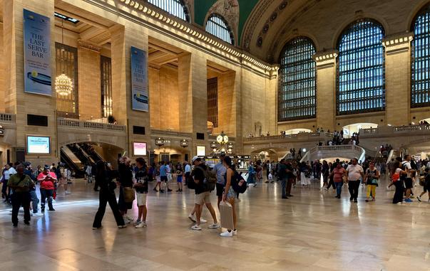 A color wide-angle photograph, an eye-level shot of the main concourse of Grand Central Terminal in New York City. The large, open room is filled with people walking in different directions. The walls and arched ceilings are made of beige and yellow-toned stone. A large clock is visible in the center of the frame. Sunlight streams in through the three tall arched windows on the far wall. Below the windows the Apple logo that adorns the elevated Apple retail store is visible. On the left side, large banners hang from the upper level. Between the columns very large, brightly lit chandeliers in the walkway outside the concourse are visible. The floor is a polished light-colored stone, reflecting the warm light and the architectural details above.