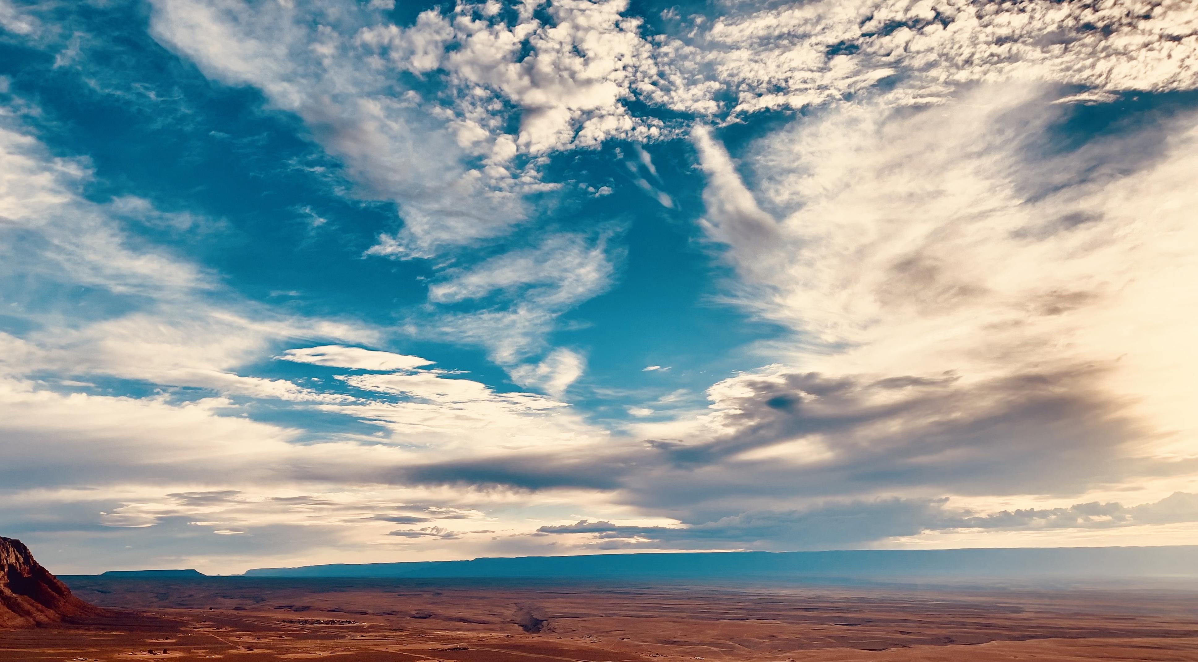 A color photograph, a landscape shot of a vast desert plain under a dramatic, multicolored sky in the late afternoon. In the foreground and middle ground, the terrain is flat, a dusty, reddish-brown expanse with a few scattered bushes and rocky outcroppings. A few small homes and trailers are seen as tiny dots across the sweeping terrain. The sky dominates the upper two-thirds of the image. The clouds are a mix of bright white, darker gray, and streaks of wispy cirrus, painted across a deep blue sky that fades to a warm, sun-kissed yellow and orange along the horizon. A distant, hazy series of plateaus forms a dark, thin line across the horizon.