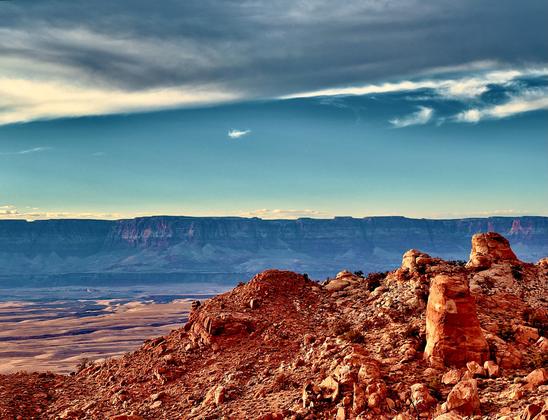 A horizontal landscape photograph taken in the late afternoon from a high vantage point overlooking Marble Canyon in Arizona. The scene is dominated by layers of geologic formations under a vast sky. In the foreground, large, rust-colored boulders and rock outcroppings, a mix of smooth and jagged textures, are illuminated by warm, directional light, casting soft shadows. The midground is a wide, flat desert landscape with a light tan and brown color palette. The background is defined by immense, dark blue and purple cliffs. The cliffs are topped with a lighter band of reddish rock, catching the last rays of sunlight. Above the landscape, the sky occupies the top half of the frame. The lower sky is a clear, light blue that transitions to a deeper blue. A long, thin cloud stretches horizontally across the middle of the sky, with a few wispy white clouds scattered above it, all set against a dramatic, dark gray and deep blue cloud layer at the very top.