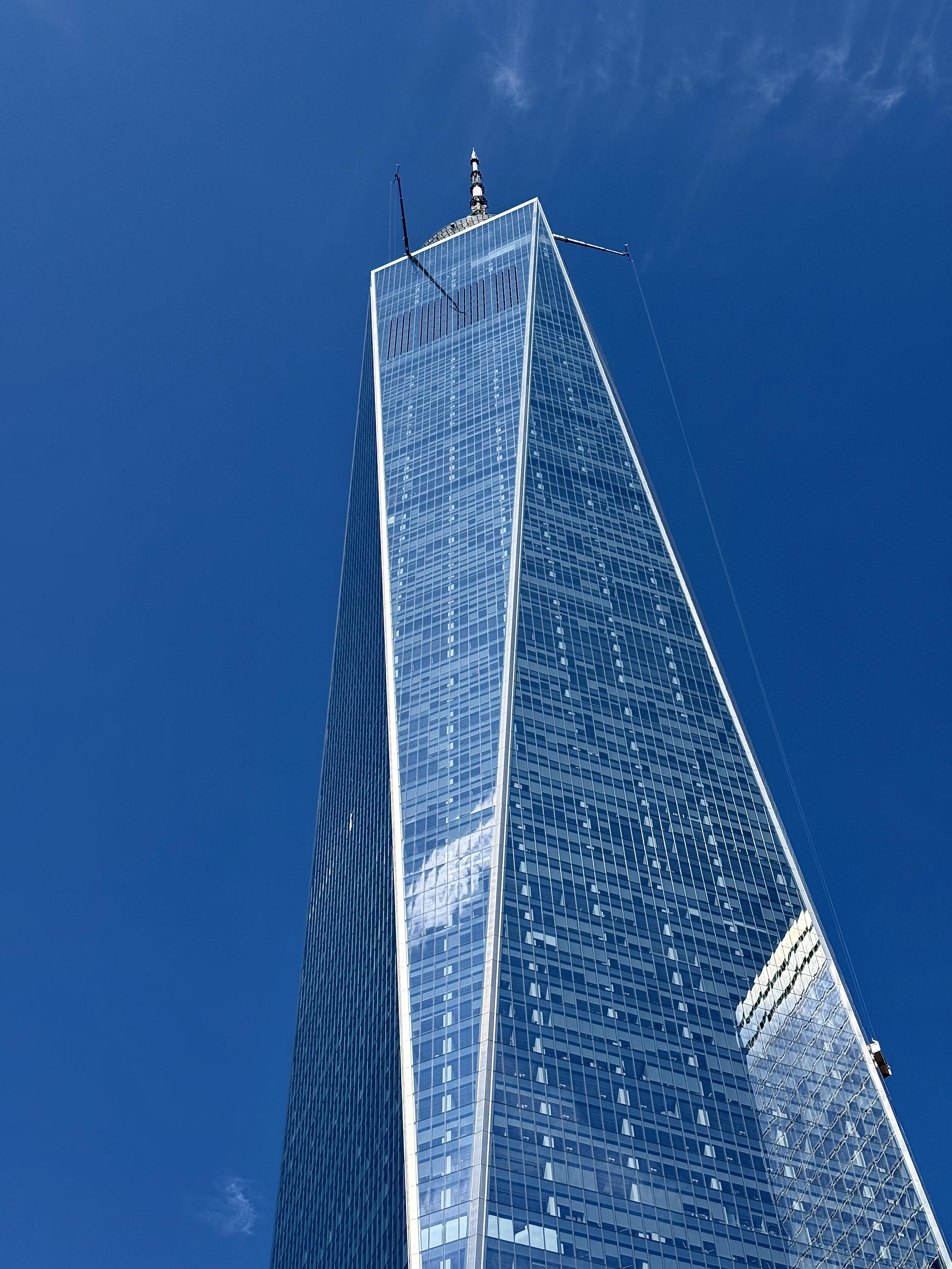 A color photograph in portrait orientation of the upper 2/3rds of One World Trade Center. The sky is a deep blue, and clear besides one wispy cloud in the upper right corner of the frame. Nearby buildings are reflecting off the glass windows. Two cranes are extending off either side of the building and a window cleaner is being suspended off the side of the building.
