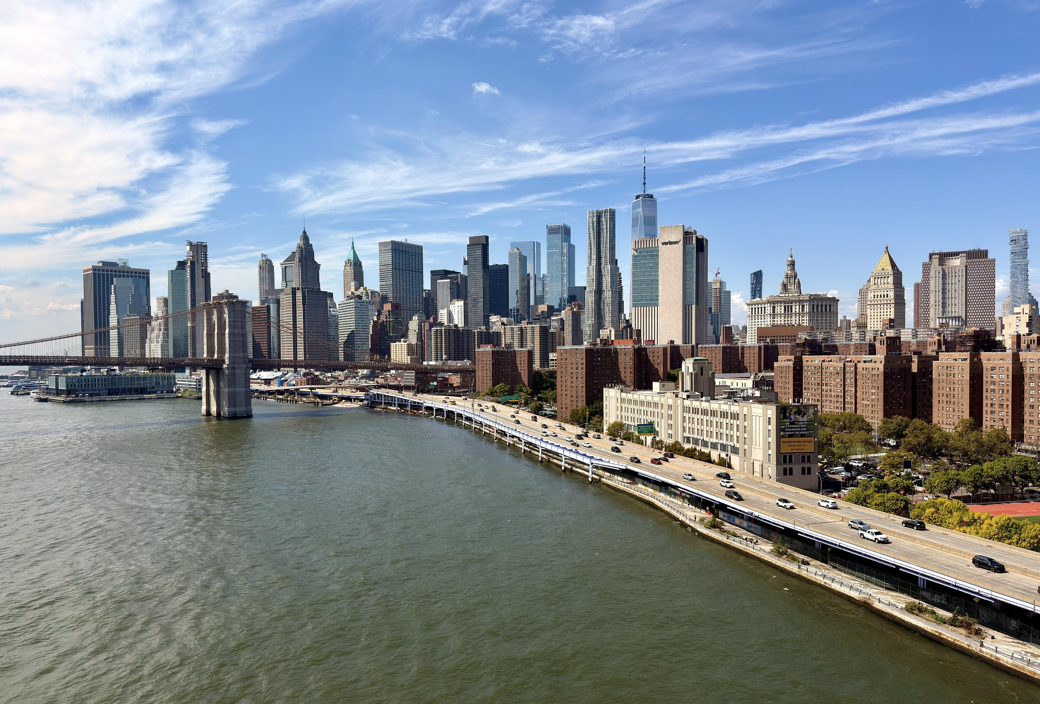 A color photograph, a panoramic, eye-level shot taken from the pedestrian walkway on the Manhattan bridge captures the lower Manhattan skyline on a sunny day. The East River, with its calm, green-brown water, fills the foreground. In the mid-ground, the Brooklyn Bridge arches gracefully from the left, its two iconic stone towers and intricate cable network visible. A steady stream of traffic moves along the FDR Drive on the right side of the frame, which parallels the riverbank. The dense collection of skyscrapers that make up the Financial District dominates the background. The prominent One World Trade Center stands as the tallest structure in the center of the skyline. The sky is bright blue with scattered white clouds, and the sunlight creates sharp reflections on the glass facades of the skyscrapers and casts long shadows on the buildings along the river.