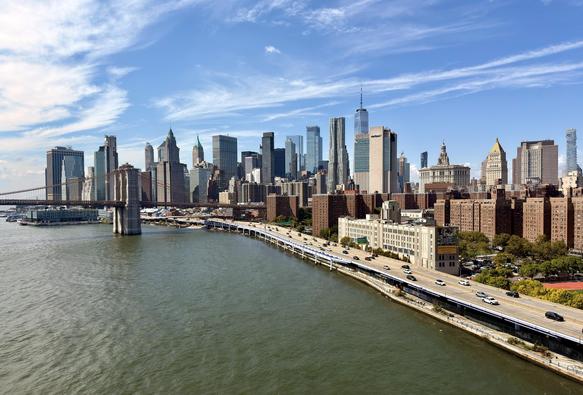 A color photograph, a panoramic, eye-level shot taken from the pedestrian walkway on the Manhattan bridge captures the lower Manhattan skyline on a sunny day. The East River, with its calm, green-brown water, fills the foreground. In the mid-ground, the Brooklyn Bridge arches gracefully from the left, its two iconic stone towers and intricate cable network visible. A steady stream of traffic moves along the FDR Drive on the right side of the frame, which parallels the riverbank. The dense collection of skyscrapers that make up the Financial District dominates the background. The prominent One World Trade Center stands as the tallest structure in the center of the skyline. The sky is bright blue with scattered white clouds, and the sunlight creates sharp reflections on the glass facades of the skyscrapers and casts long shadows on the buildings along the river.