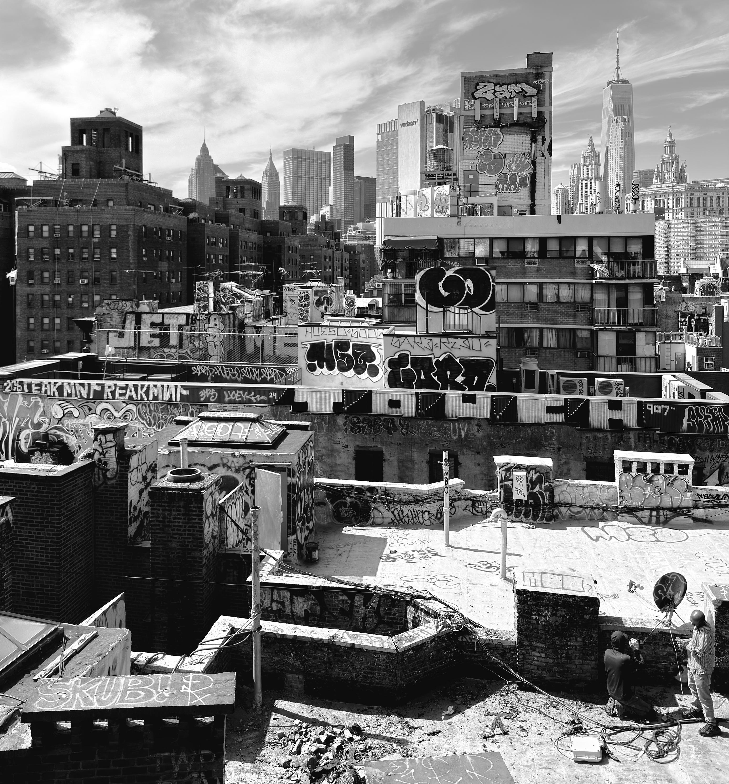 A black-and-white photograph of a rooftop view in the Two Bridges neighborhood of lower Manhattan on a bright day. The foreground shows many cluttered roofs covered in rubble and various vent pipes and brick enclosures. The walls and many of the rooftop structures are heavily covered in graffiti tags. In the bottom right corner, two individuals are working. One person, wearing a hoodie and shorts, kneels on the rooftop, while another person stands beside them. Wires and equipment are scattered on the ground around them. The skyline is filled with a variety of skyscrapers under a partly cloudy sky, most notably One World Trade Center which towers above the rest.